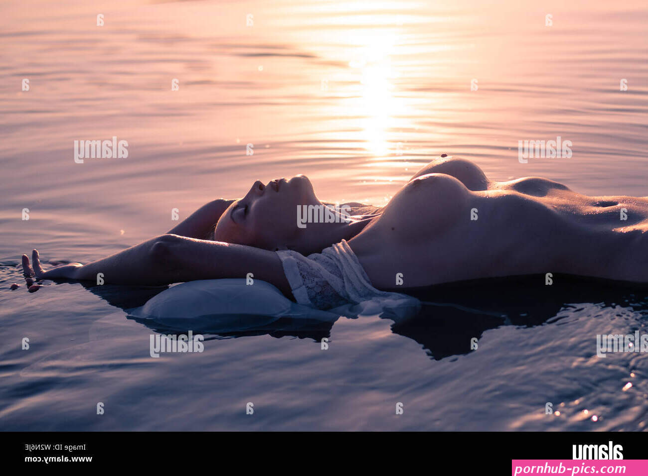 Silhouette of a young sexy nude woman with big beautiful breasts lies in the waves of the reservoir in the rays of the setting sun Stock Photo