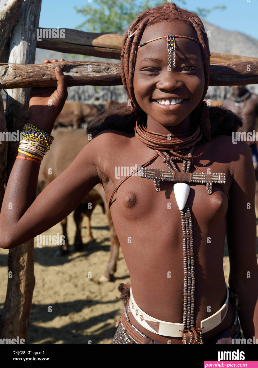 Namibia Kaokoland. A happy young Himba girl at the entrance to her familys stock pen. Her body gleams from a mixture of red oc Stock Photo