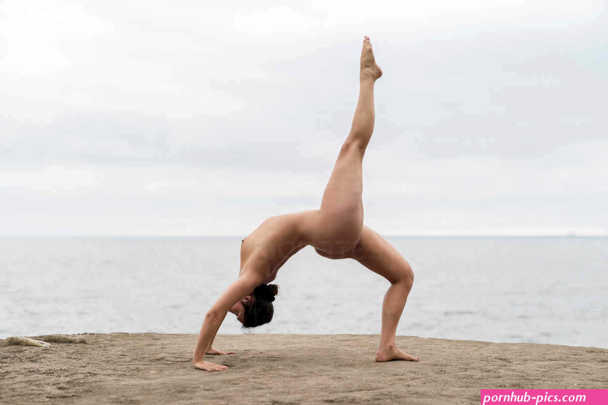 Flexible young naked woman practicing bending over backwards at beach against sky stock photo