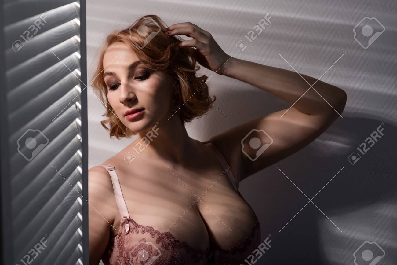 A beautiful young woman with very large natural breasts is standing by the window with shutters. A girl in pink lace shows a beautiful figure next to a white screen. Huge female