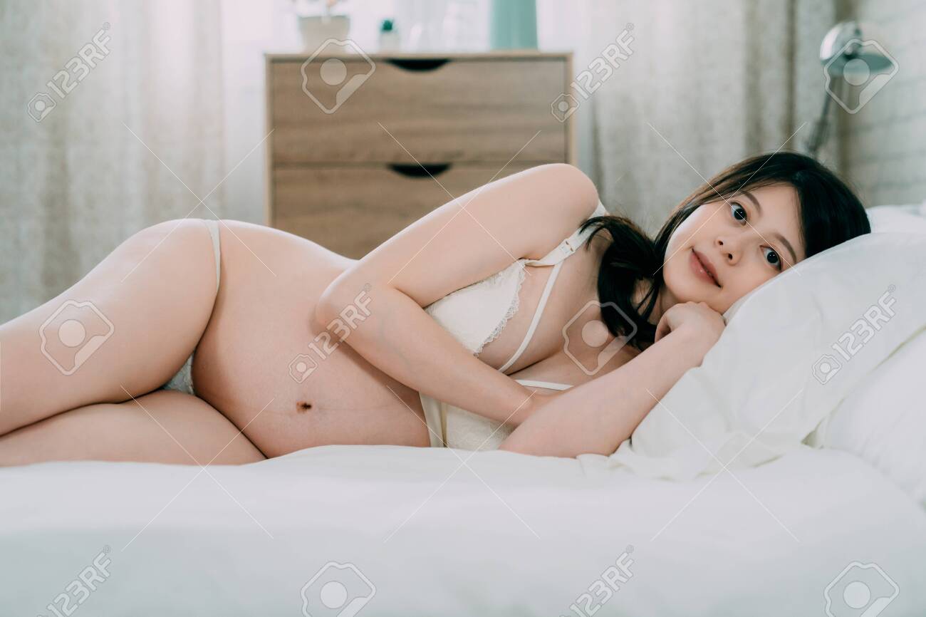 Beautiful Sexy Pregnant Woman Lying On Bed In Bedroom At Home. Elegant Young Mom With Bare Belly Dressed In White Fashion Lingerie. Charming Maternity Lady Face Camera With Attractive Smile. Stock Photo